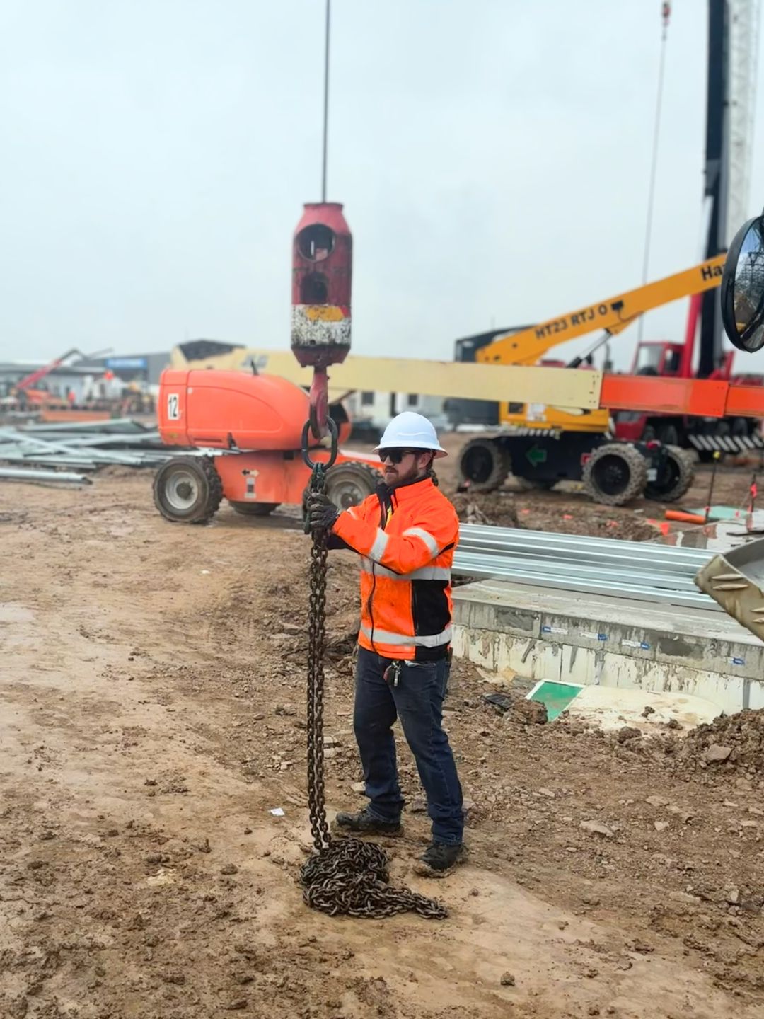 An SLE worker in hi-vis and hard hat on a live construction site, checking a lifting chain connected to a crane hook, with cranes and a boom lift behind